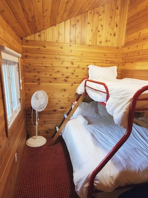 A wooden cabin room with bunk beds, a standing fan, and a window with blinds.