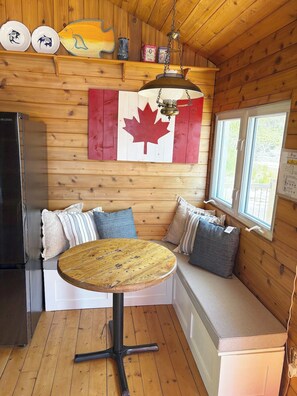 A cabin interior with a wooden interior, a dining table, a bench with cushions, a refrigerator, and a window with a view.