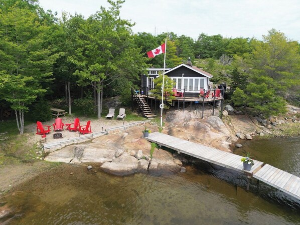 A lakeside cabin with a dock, red chairs, and a Canadian flag.