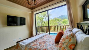 Une chambre à coucher avec un lit, un téléviseur fixé au mur et un balcon offrant une vue sur les arbres et les montagnes.
