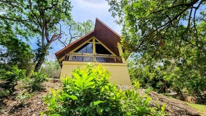 Un petit bâtiment jaune, avec un toit brun et un balcon, entouré de verdure et d’arbres.