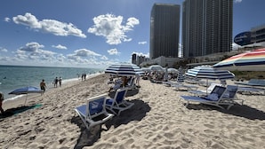 A beach with sun loungers and umbrellas, a clear sky, and tall buildings in the background. - Exclusive 3 bedrooms apartment w Amazing View (Hallandale Beach)