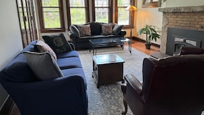 A living room with a sofa, armchair, and coffee table, a fireplace, and large windows with a view of a garden. - Walk to Beach Lake of Michigan, Lake Park, Eastside Milwaukee, Sleep 6, Parking (Milwaukee)