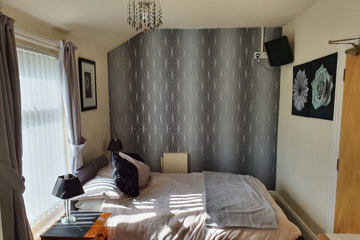 Ein Schlafzimmer mit einem Bett, einem Nachttisch, einer Lampe, einem an der Wand montierten Fernseher und einem Fenster mit Vorhängen.