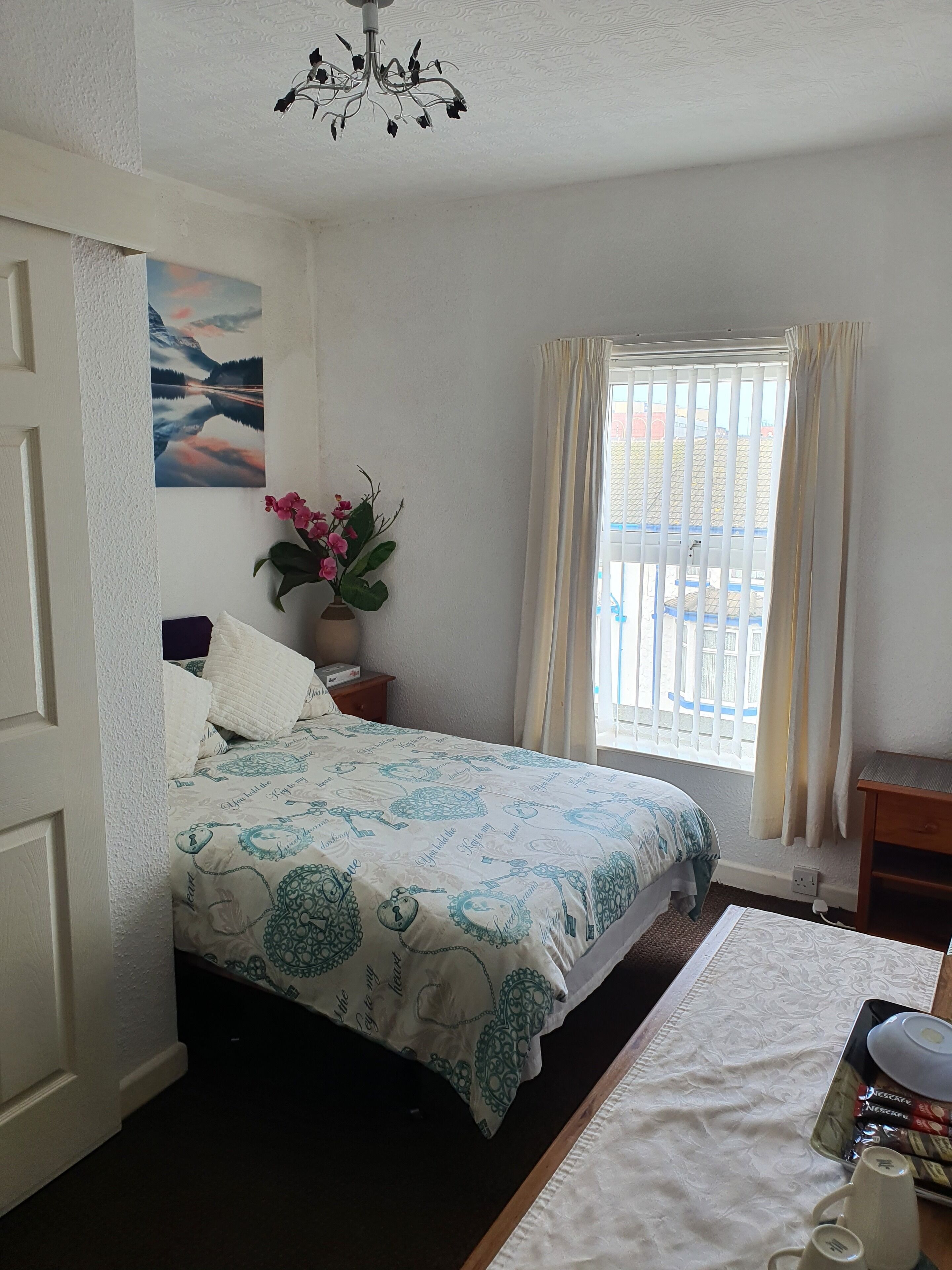 A bedroom with a bed, a nightstand, a vase with flowers, a window with curtains, and a painting on the wall.
