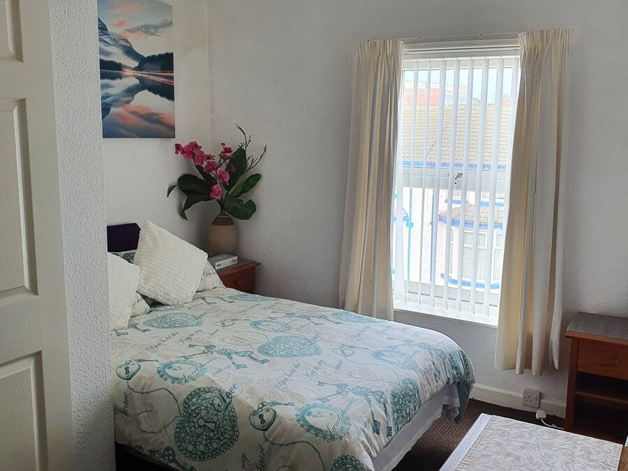 A bedroom with a bed, a nightstand, a vase with flowers, a window with curtains, and a painting on the wall.