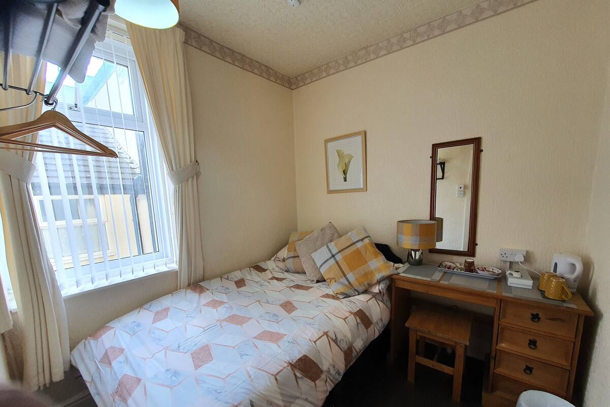 Ein Schlafzimmer mit Bett, Schreibtisch, Stuhl, Spiegel, einer Pendelleuchte und einem Fenster mit Vorhängen.