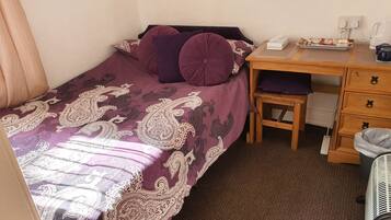 Un dormitorio pequeño y ordenado con una cama individual, un escritorio de madera, una silla y una mesita con una caja de pañuelos y una tetera.