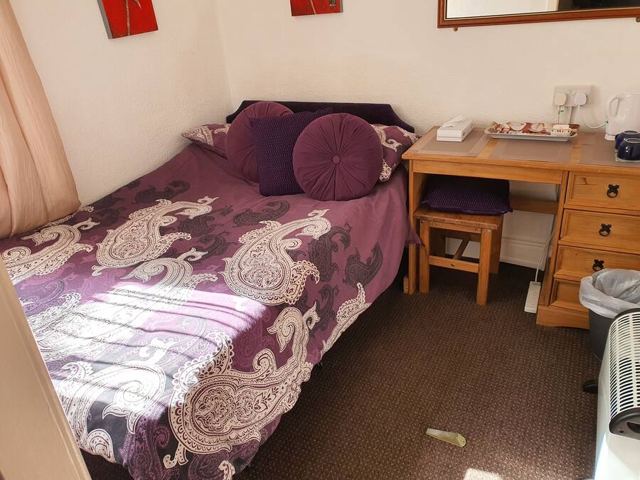 Un dormitorio pequeño y ordenado con una cama individual, un escritorio de madera, una silla y una mesita con una caja de pañuelos y una tetera.