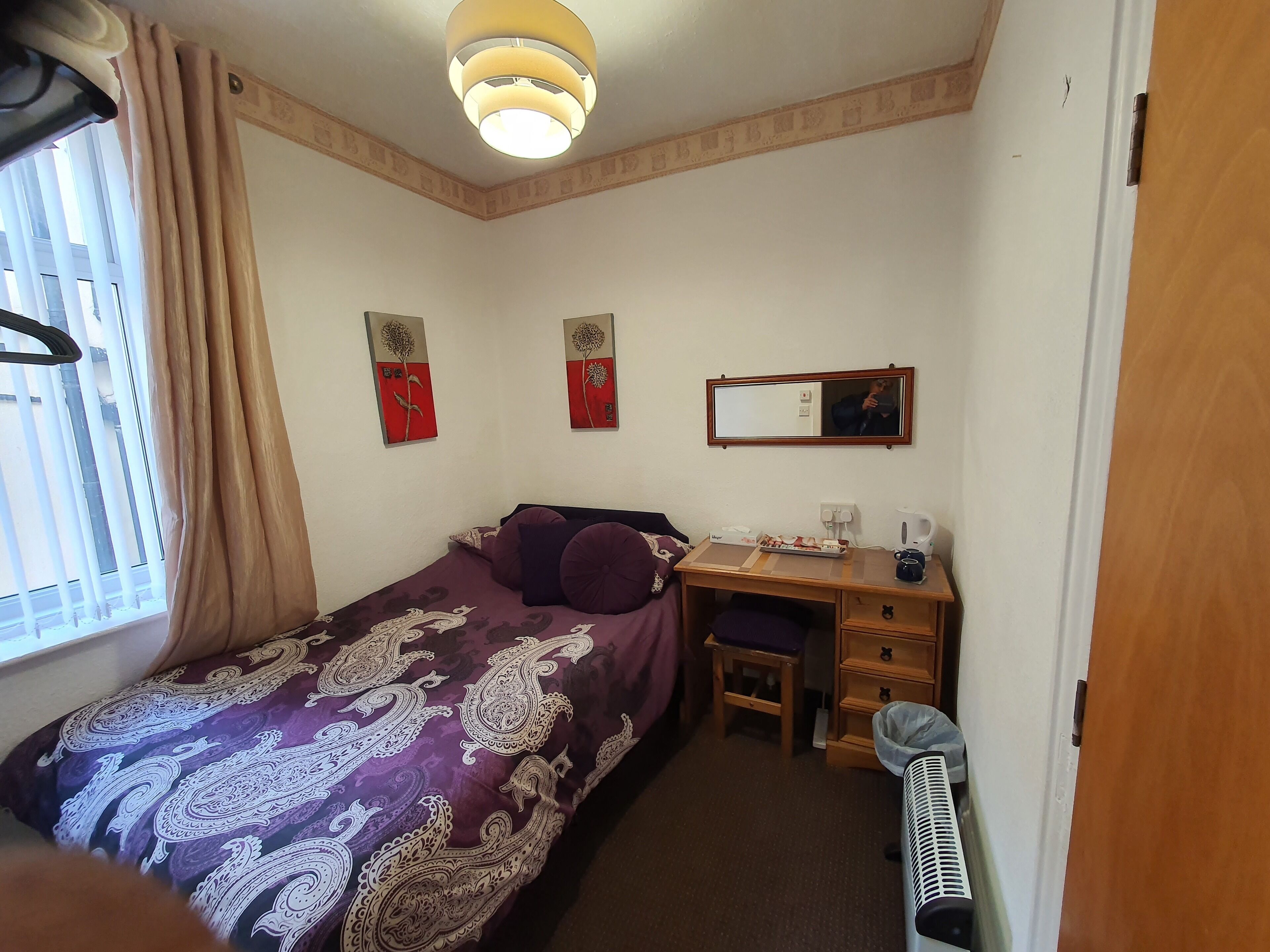 Habitación de hotel pequeña con cama, escritorio, silla, mesita, radiador y ventana con cortinas.