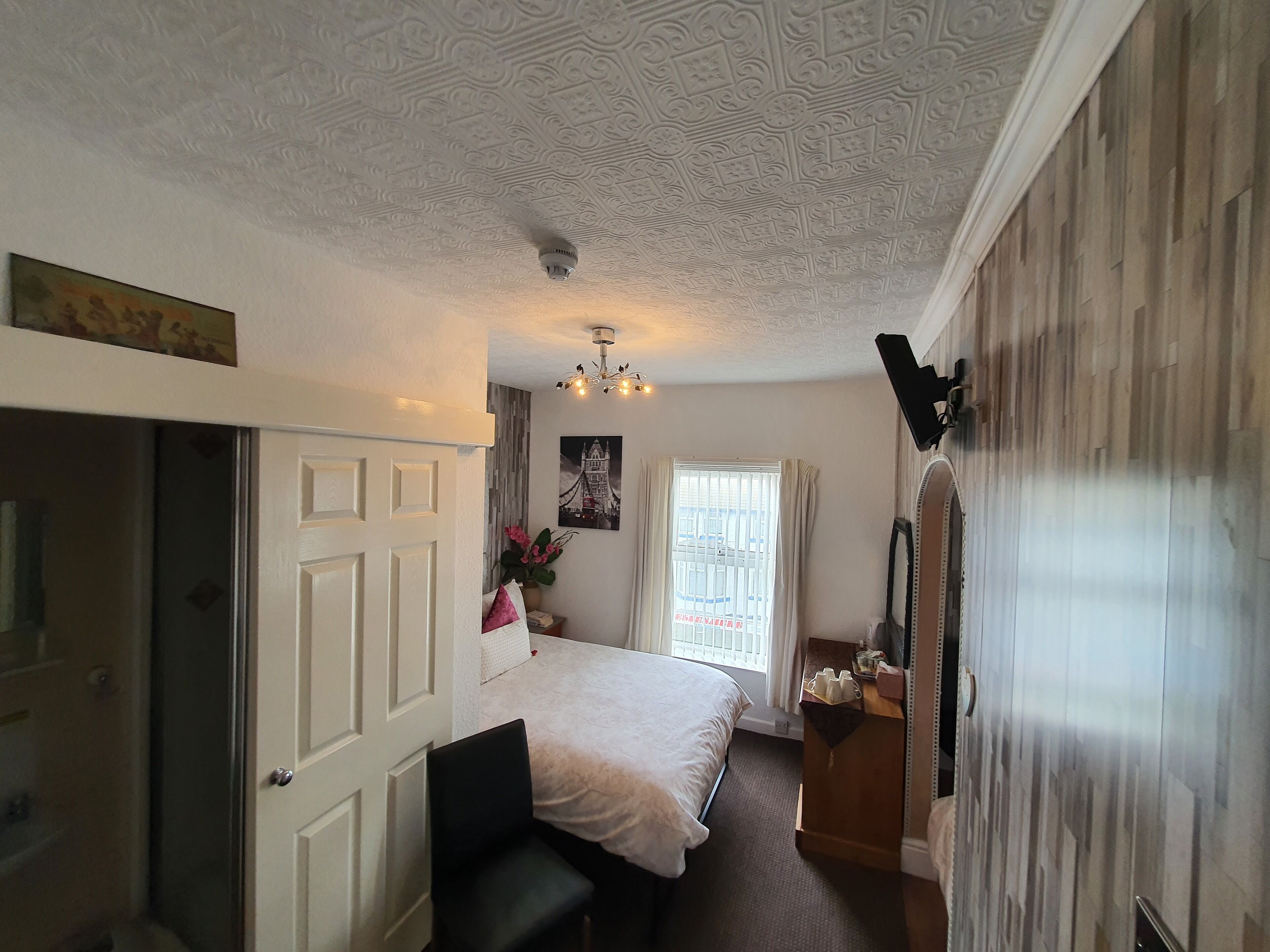 A bedroom with a bed, a desk, a chair, a window with curtains, a ceiling with intricate patterns, and a wall-mounted light fixture.