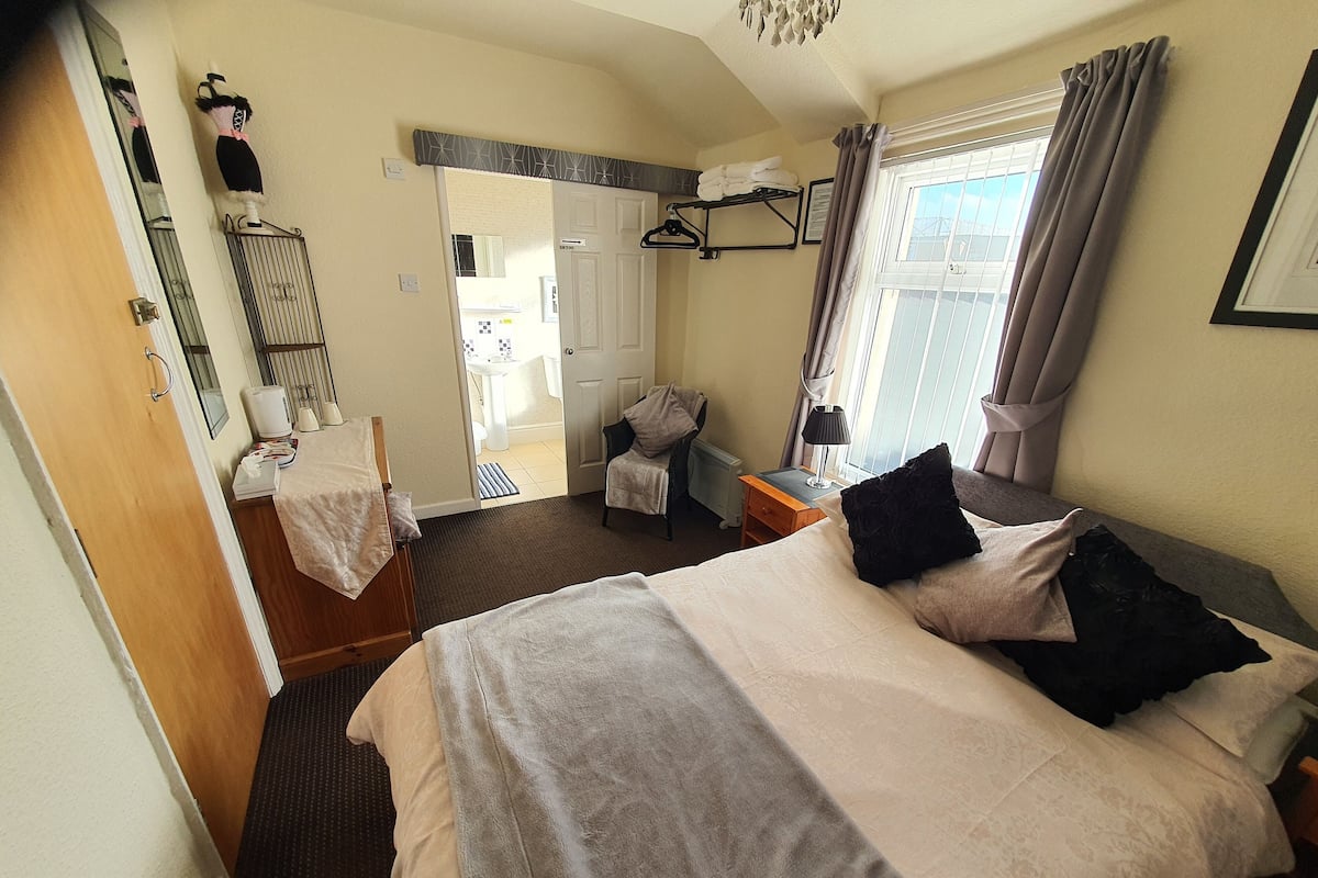 Ein Hotelzimmer mit Bett, Stuhl, Schreibtisch, Spiegel, einem Fenster mit Vorhängen und einer Tür zu einem Badezimmer.