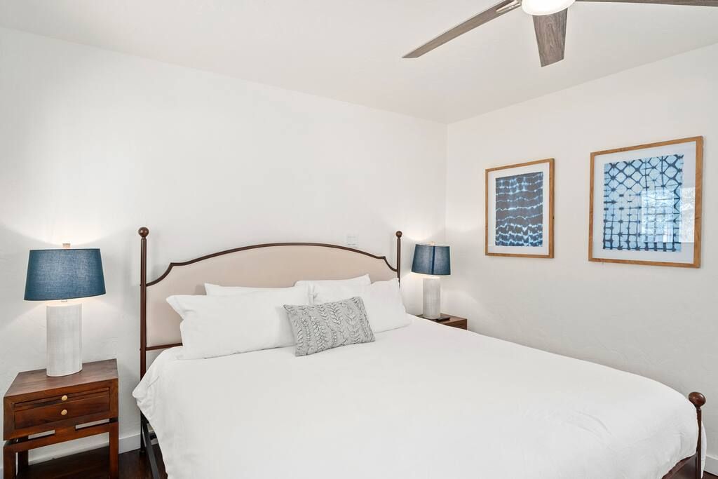 A neatly made bed with white linens and a gray pillow, flanked by two bedside tables with lamps, in a room with white walls and two framed artworks.