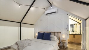 A bedroom with a bed, bedside tables, a nightstand, a small table, and a mirror. There is a kitchen area visible through an open doorway.
