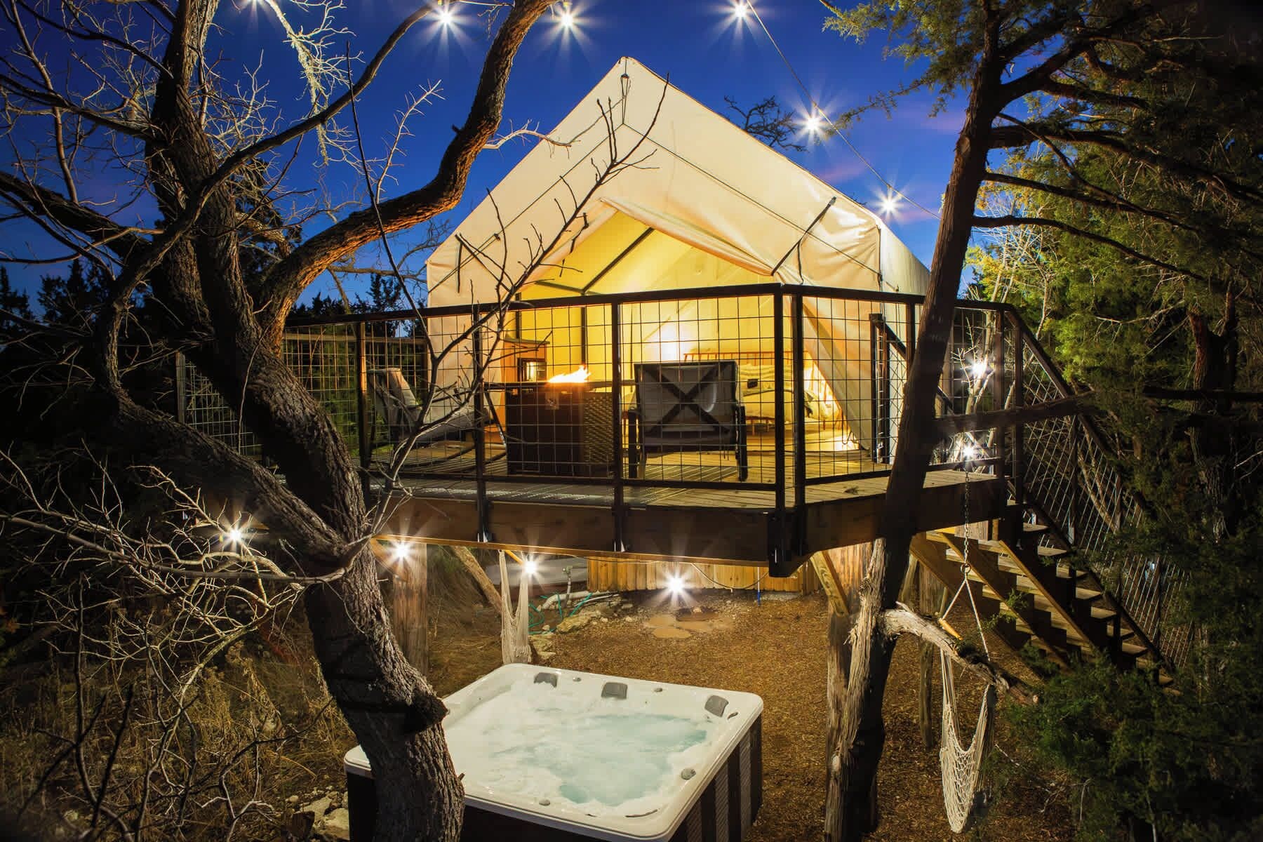 A treehouse with a hot tub, surrounded by trees and a tent-like structure.