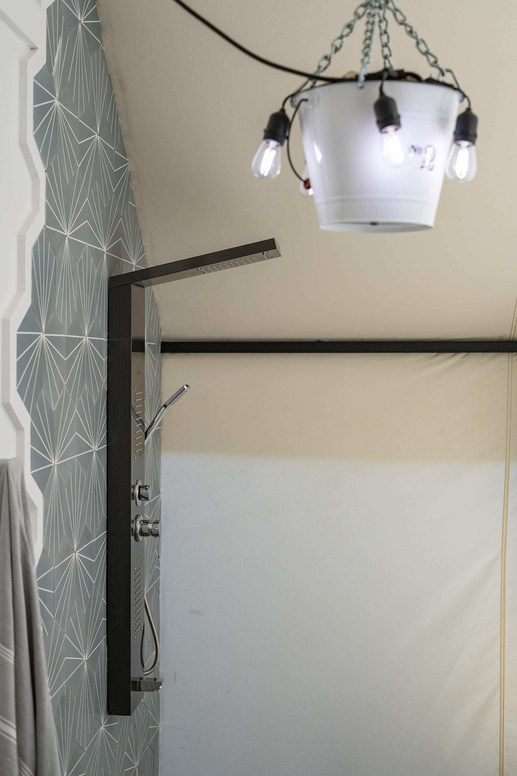 Een badkamer met een geometrisch patroon op de muur, een douche met een handdouche en een hangende lamp met zichtbare gloeilampen.