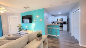 A modern living room with a teal accent wall, a flat-screen TV, and a kitchen area with white cabinets and stainless steel appliances.