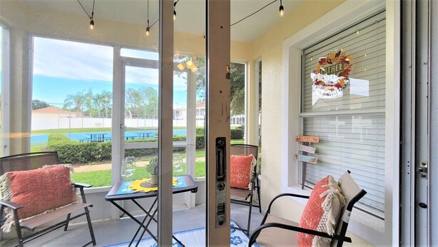 A covered patio area with two chairs, a small table, and a view of a swimming pool and tennis courts.