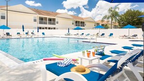 A pool area with lounge chairs and umbrellas, a poolside table with drinks, and a building in the background.