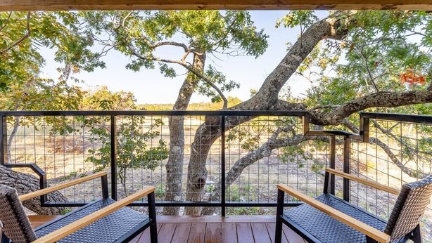 Une terrasse en bois, agrémentée de deux fauteuils en osier et d’une rambarde en métal, qui offre une vue sur un paysage naturel avec des arbres et un ciel dégagé.