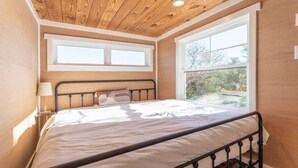 Une chambre avec un plafond en bois, un lit avec un cadre en métal, deux fenêtres et une vue sur les arbres.