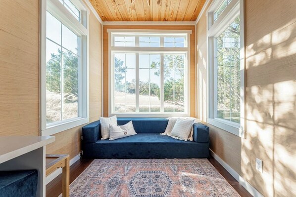 Ein Zimmer mit einem blauen Sofa, einem gemusterten Teppich und großen Fenstern mit Blick auf Bäume und einen klaren Himmel.