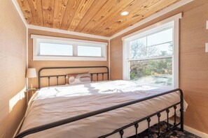 Ein Schlafzimmer mit Holzbalkendecke, einem Bett mit Metallgestell, zwei Fenstern und Blick auf Bäume.