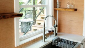 Une cuisine avec une fenêtre, un évier en acier inoxydable et des étagères en bois où sont disposés des bocaux en verre et des verres à vin.