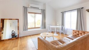 Ein geräumiges Wohnzimmer mit einer Ledersofa, einem runden Couchtisch und einem Esstisch mit Stühlen. Große Fenster mit Vorhängen, eine Klimaanlage und ein Spiegel an der Wand sind ebenfalls vorhanden.