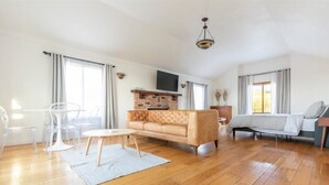 Ein geräumiges Wohnzimmer mit Holzboden, einem beigefarbenen Sofa, einem Couchtisch und einem an der Wand befestigten Fernseher.