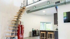 A spacious living area with a wooden staircase, a kitchen with a bar, and a large window. - Restored Grain Bin with Hot Tub, King Bed, Vending Machine. Dog-friendly! (Rockbridge)