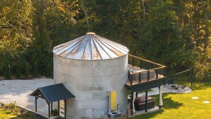 A cylindrical metal structure with a balcony, surrounded by a grassy area and trees. - Restored Grain Bin with Hot Tub, King Bed, Vending Machine. Dog-friendly! (Rockbridge)