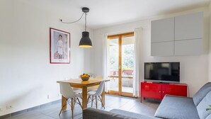 A living room with a sofa, a dining table with chairs, a TV on a red cabinet, and a large window with curtains.