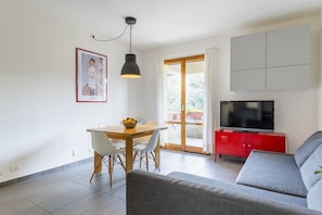 Un salon comprenant un canapé, une table à manger avec des chaises, un téléviseur posé sur un meuble rouge et une grande fenêtre avec des rideaux.