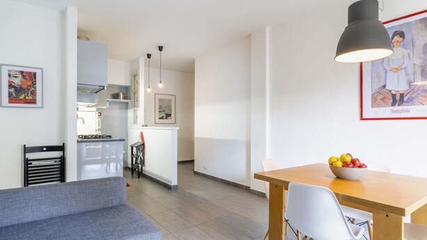 A modern living room with a grey sofa, a wooden dining table with white chairs, a kitchen in the background, and a pendant light.
