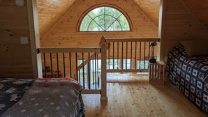 A cozy cabin with a loft bedroom, a large arched window, and a wooden ceiling.