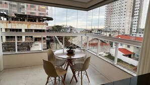 A modern dining area with a round table and chairs, large windows offering a view of a construction site and surrounding buildings. - Ap Gold Coast Vista Mar - Piçarras - SC (Picarras)