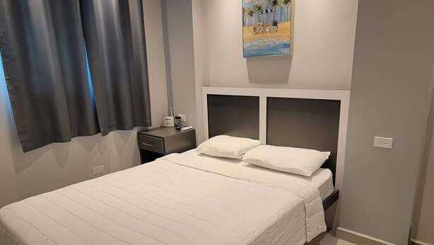 A neatly made bed with a quilted cover, a bedside table with a lamp, and a painting above the bed. - SUITE 203 - 2 PEOPLE (Cabo Rojo)