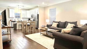 A modern living room with a sofa, coffee table, and TV. There is a kitchen area with cabinets and a dining table. - Luxury Townhouse Near Lakefront & Downtown (Lake Charles)