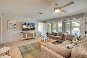 A living room with a beige sofa, a television, a ceiling fan, and a view of the outdoors through large windows. - Simpatico (Anna Maria)