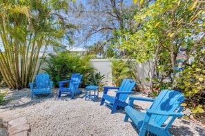 A patio with blue Adirondack chairs and a gravel ground, surrounded by various plants and trees. - Simpatico (Anna Maria)