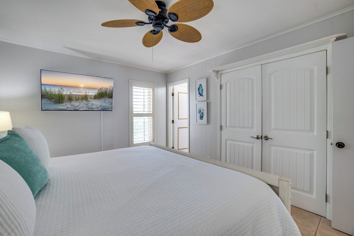 A bedroom with a large bed, a ceiling fan, a painting of a beach scene, and white doors.