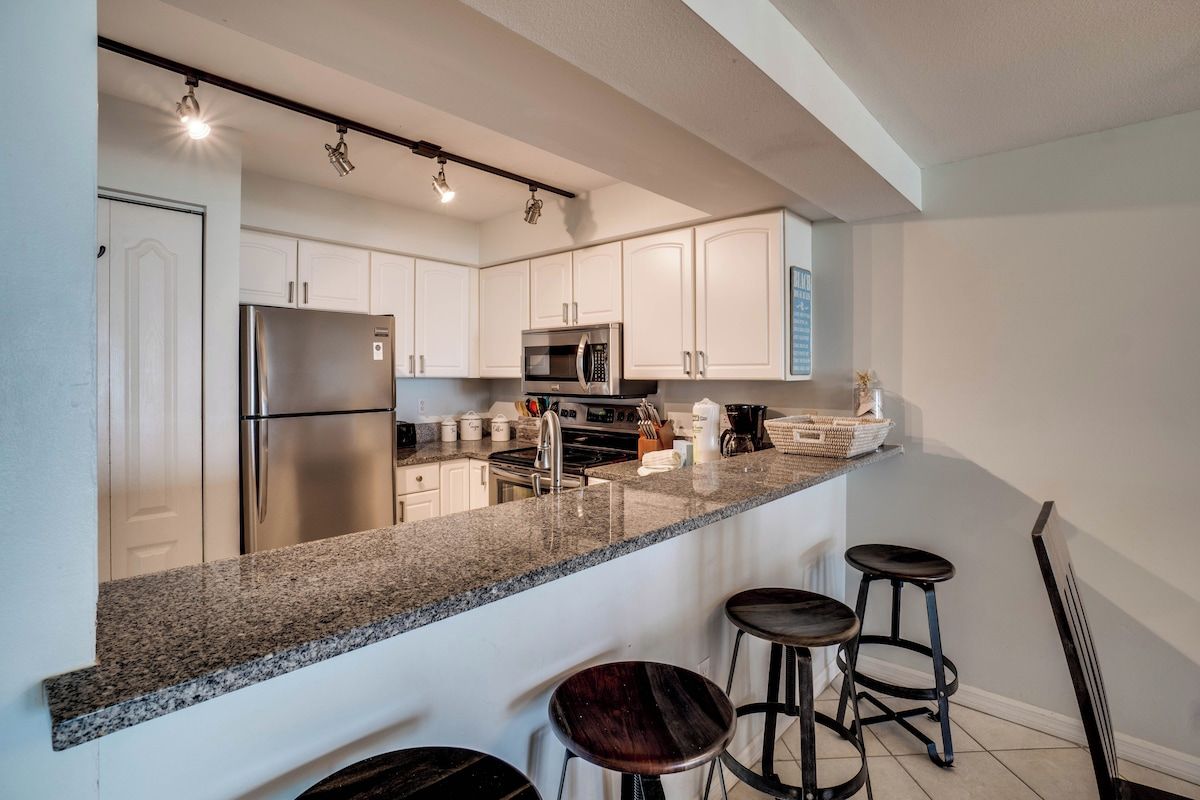 Et moderne køkken med stålemmer, lyse skabe og en bordplade i granit. Tre barstole og en lille ø med vask.