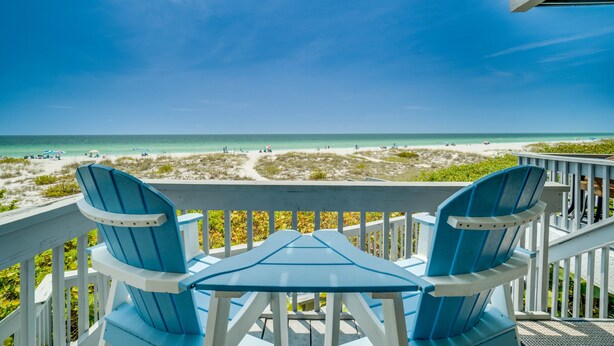 Een balkon met twee blauwe stoelen en een tafel, met uitzicht op een strand met mensen en parasols.