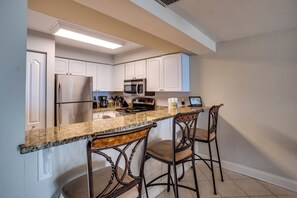 Una cocina con gabinetes blancos, electrodomésticos de acero inoxidable, encimera de granito y dos taburetes de bar.