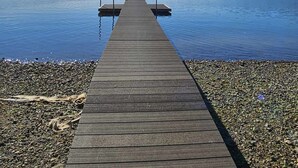 Een houten steiger die zich uitstrekt over kalm water, omringd door een kiezelstrand en een bebost achtergrond.
