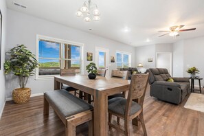 A modern living room with a dining area, a ceiling fan, and large windows with blinds. - Bright Open Floorplan with Hot Tub, Private Deck, Arcade, Golf Course & High-End Decor (Branson West)