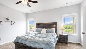 A bedroom with a bed, two bedside tables, a ceiling fan, and two windows with a view of greenery. - Bright Upgraded Decor with Hot Tub, Deck, Arcade, Free Tickets & Onsite Ledgestone Golf (Branson West)