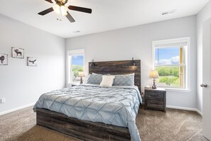 A bedroom with a bed, two bedside tables, a ceiling fan, and two windows with a view of greenery. - Bright Upgraded Decor with Hot Tub, Deck, Arcade, Free Tickets & Onsite Ledgestone Golf (Branson West)