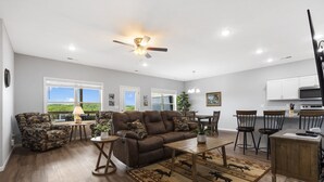 A spacious living room with a brown leather sofa, a coffee table, and a patterned rug. There are multiple windows offering a view of the outdoors. - Bright Upgraded Decor with Hot Tub, Deck, Arcade, Free Tickets & Onsite Ledgestone Golf (Branson West)
